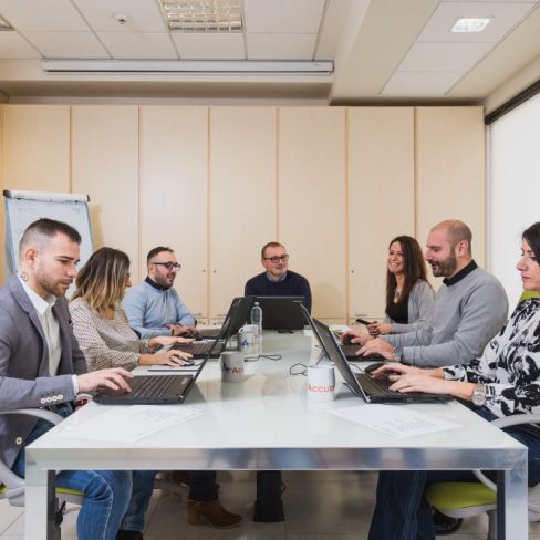 Gruppo di persone sedute ad un tavolo che lavorano assieme. Ogni persona ha davanti a se un computer portatile e una tazza con la scritta 'Accueil'.