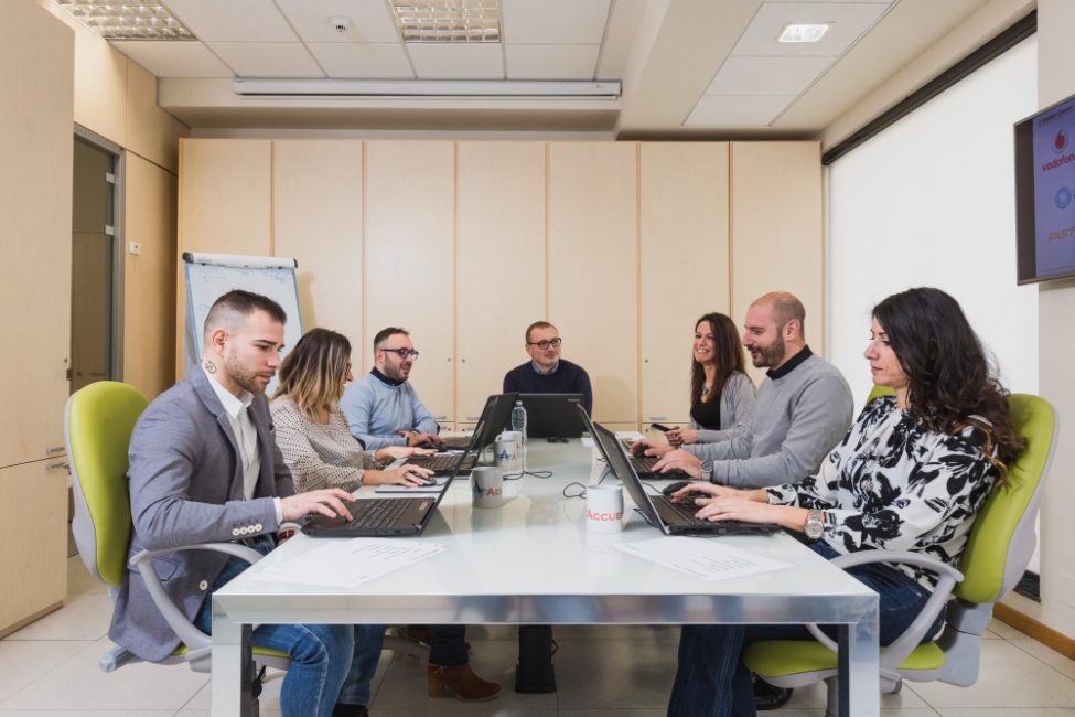Gruppo di persone sedute ad un tavolo che lavorano assieme. Ogni persona ha davanti a se un computer portatile e una tazza con la scritta 'Accueil'.