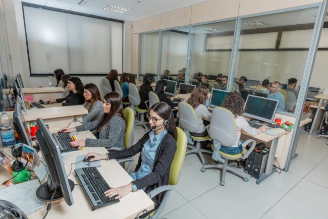 Gruppo di persone sedute alle proprie postazioni che indossano delle cuffie e lavorano al computer.