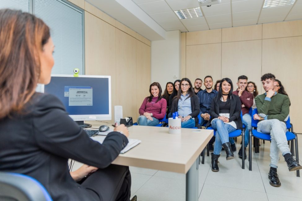Gruppo di persone sedute in una stanza che ascoltano un'interlocutrice sedute alla scrivania.