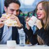 Uomo e donna seduti ad un tavolo. L'uomo tiene in mano un panino mentre la donna tiene in mano una tazza con la scritta 'Accueil'.