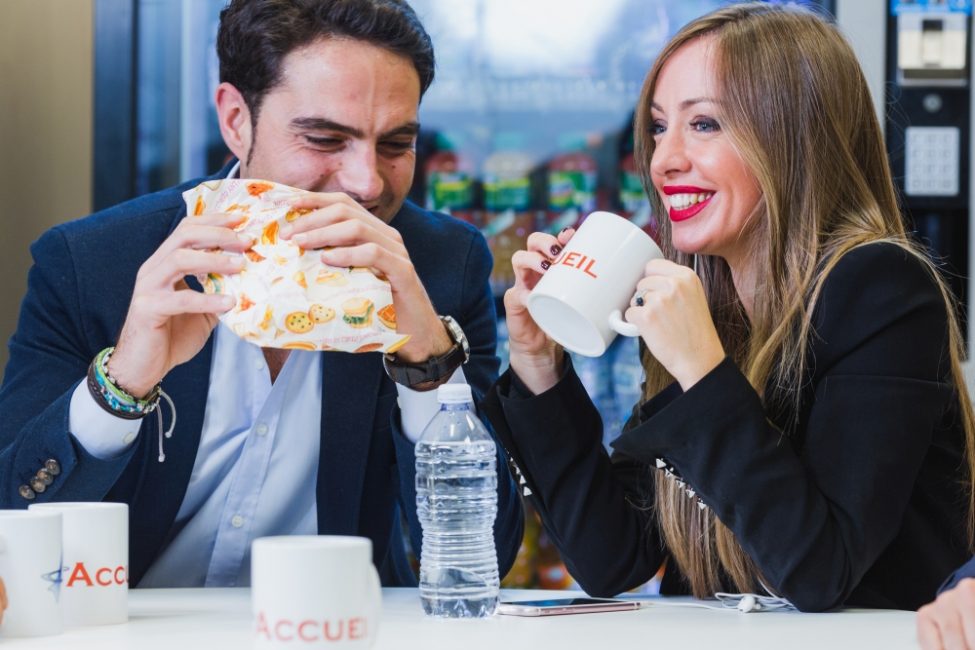 Uomo e donna seduti ad un tavolo. L'uomo tiene in mano un panino mentre la donna tiene in mano una tazza con la scritta 'Accueil'.