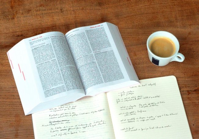 Dizionario, quaderno e tazzina con il caffè appoggiati su un tavolo.