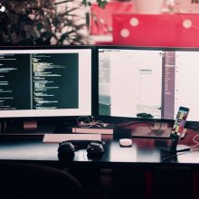 Una scrivania con due schermi di computer, delle cuffie e un cellulare.