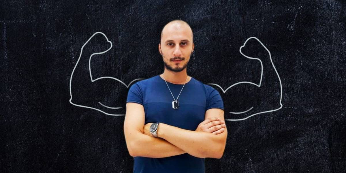 Uomo con le braccia conserte. Sullo sfondo vi è una lavagna con disegnate delle braccia muscolose.