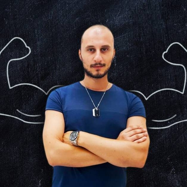 Antonino-Calarco Uomo con le braccia conserte. Sullo sfondo vi è una lavagna con disegnate delle braccia muscolose.