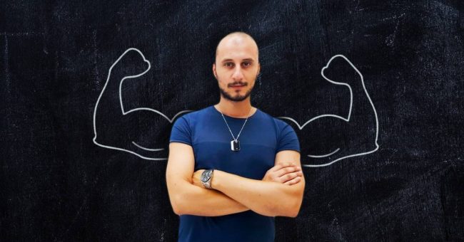 Antonino-Calarco Uomo con le braccia conserte. Sullo sfondo vi è una lavagna con disegnate delle braccia muscolose.