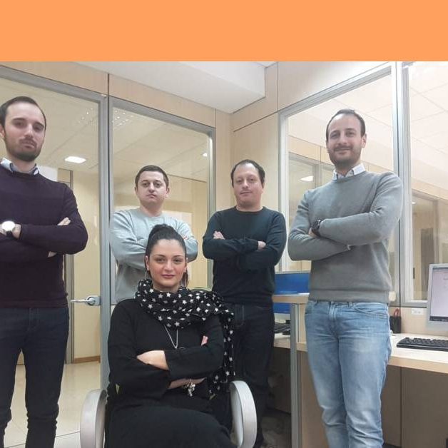 Antonella-Lagana Gruppo di persone in piedi con le mani conserte e una persona seduta su una sedia.