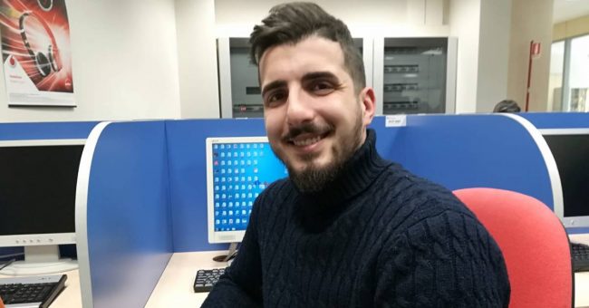 Ragazzo in ufficio che sorride seduto su una sedia. Sullo sfondo una postazione con il computer.