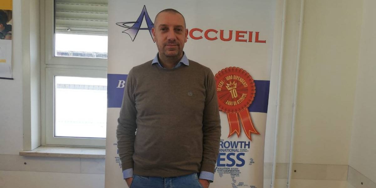 Dario-Sanguedolce Uomo con le mani in tasca in piedi davanti a uno striscione con il logo dell'azienda Accueil