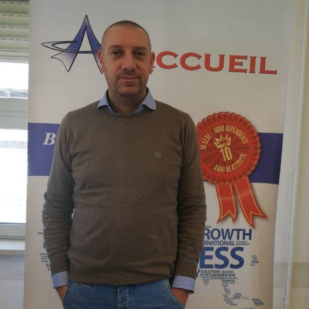 Dario-Sanguedolce Uomo con le mani in tasca in piedi davanti a uno striscione con il logo dell'azienda Accueil