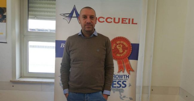 Uomo con le mani in tasca in piedi davanti a uno striscione con il logo dell'azienda Accueil