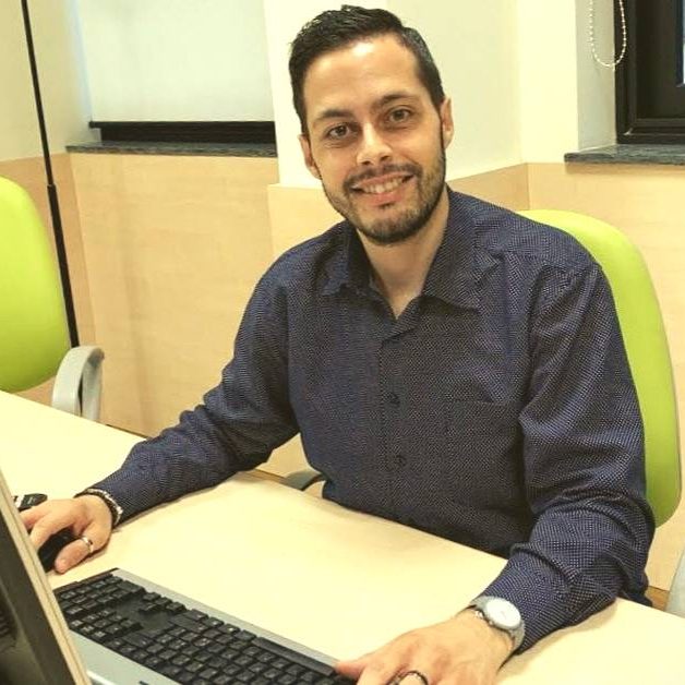 Roberto-Pugliatti Uomo che sta sorridendo in ufficio ed è seduto su una sedia verde di fronte al computer