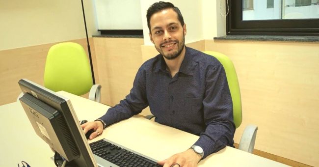Uomo che sta sorridendo in ufficio ed è seduto su una sedia verde di fronte al computer