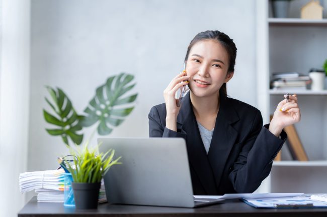 Ragazza che parla al telefono davanti ad un computer portatile e tiene in mano una penna.